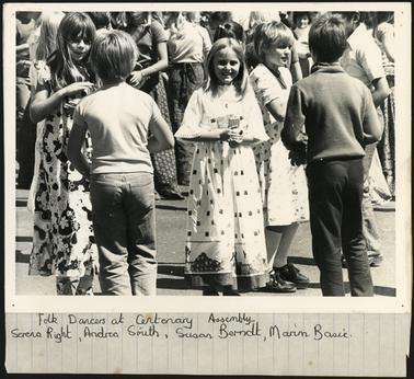 This image shows several children dancing. In clear view are 3 girls and 2 boys. The girls are facing the camera, and the boys have their backs to the camera. In the background are some adults, they may be dancing too. 