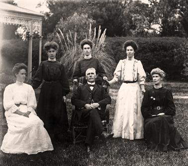 A family group of people pictured in a garden near the corner of a verandah. An elderly man is seated between an older and a younger woman with three young women standing at the rear. The verandah has an iron lace frieze and the figures are in front of some shrubs in a neat garden which is bordered by a hedge.