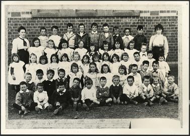 This class photo shows 55 pupils, girls and boys. These are ranged in 5 tiers. There are two teachers shown standing, one either end of the group. There is a brick wall in the background, which appears to be the school.
