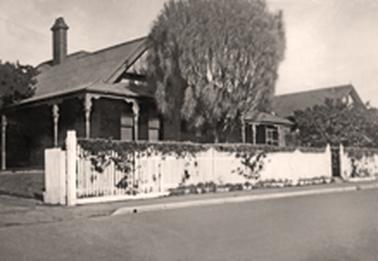 An Edwardian single storied house with verandah behind a white picket fence on the street. The house has a pitched roof with a single, tall chimney. There is a driveway on the left with open gates, a small pedestrian gateway to the right and a tall tree between house and fence. A shrub conceals what appears to be an extension of the house.