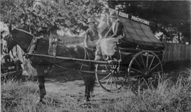 Horse and covered cart labelled Tubba Rubba Prospectors with a bucket hanging from the apex of the covering. A man and a woman are seated behind the horse and are dressed in clothes appropriate to the previous century. The horse and cart are on rough grass in front of a paling fence. The front of a car or truck, licence number 83276, is in the background to the left. 
