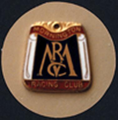 A rectangular metal badge, wider at the bottom, has a gold pattern at the top, ‘Mornington’ on a curved brown ribbon below it and ‘Racing Club’ along the brown strip at the bottom. In the centre is ’MRC’  in golden letters on a black background, with a golden whip on either side of the monogram. The badge is photographed on a fawn  coloured circular background.
