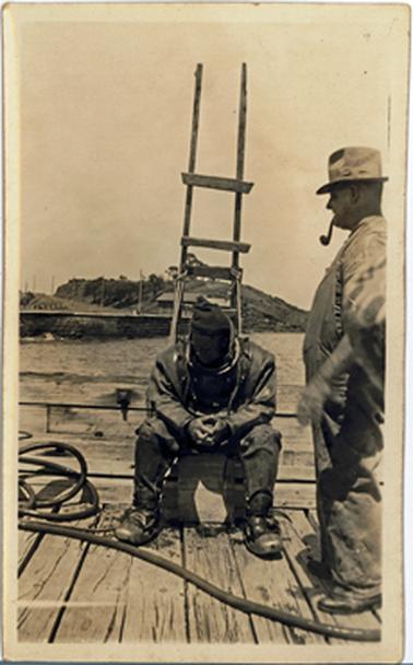 Two men on the edge of Mornington Pier. One is in diving gear with a large heavy brass and glass full head helmet and seated on a rung of a ladder which is roped to the pier. The other man, who is dressed in work clothes and a hat, is standing beside him, smoking a pipe. There is a hill in the background. 
