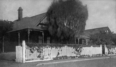 An Edwardian single storied house with verandah behind a white picket fence on the street.The house has a pitched roof with a single, tall chimney. There is a driveway on the left with open gates, a small pedestrian gateway to the right and a tall tree between house and fence. A shrub conceals what appears to be an extension of the house.