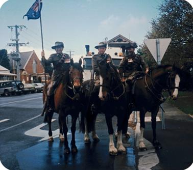 Three mounted horsemen, each wearing army uniform and slouch hats with feathers. Behind them is the truck bearing the Honour Roll they are about to escort from Elliott Square outside the Mechanics Institute and Council Chambers to Memorial Park, Mornington. St. Peter’s Anglican Church and Queen Street is in the Left background. The men are wearing numerous medals on their uniforms. One horseman is bearing a standard with the Australian flag. Two of the horses’ noses have white blazes.