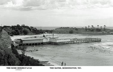 This shows the Baths from the top of the embankment. It is a cloudy day, and three people are shown in the foreground walking on the beach. Mothers Beach is shown on the far side of the Baths. There is a sign on the building saying “Welcome to Mornington, bath in safety etc”. The land on the other side of Mothers beach is covered in scrub and trees, with only a couple of sheds visible. There are several small boats on the beach in the foreground.