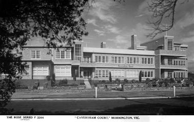 This photo shows blocks of flats. The left of the building is two storey, and to the right is three storey. The are two stairwells clearly visible. The right side of the building has a flat roof. In the centre of the photograph are steps leading up to the front entrance. There is a low stone fence across the front of the building. 
