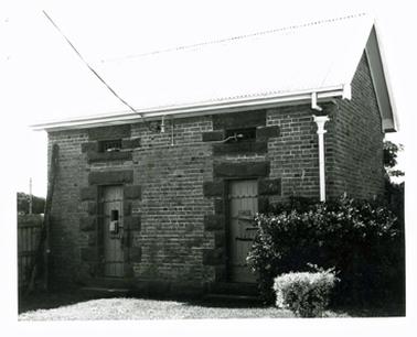 This shows a small brick building with two doors and a pitched roof. The doors appear to be re-enforced with metal bars. The roof is corrugated iron.