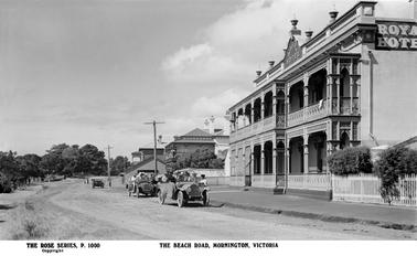 This shows the Royal Hotel on the right of the image. It is a magnificent two storey rendered building with ornate iron work to both verandahs. In the centre of the top storey is a decorative pediment showing the name of the hotel. On the roof are decorative stone urns. In the centre of the image is Kirkpatricks Hotel. There are two motor vehicles in the front of the Royal and they are full of people. There is a third vehicle parked outside Kirkpatricks, and three women walking along the road.
