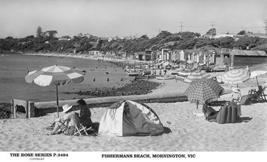 This is a lovely view showing the whole of Fishermans Beach on a summers day. It is very busy with lots of people enjoying the ambiance. There are many sun umbrellas, in the centre foreground is a sun “igloo” or tent. There are over 20 beach huts shown, and several boats on the beach edge. On the top of the embankment behind the beach and several houses, and telegraph poles.