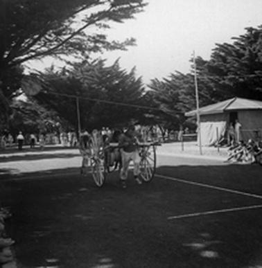 A fire hose reel is on the Mornington Park training track. It is being pulled along by a man in harness between the wheels. There are cypress trees in the background and onlookers are gathered in groups beside the track.