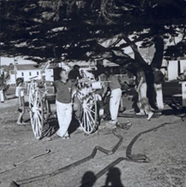 An empty fire hose reel sits under a tree canopy with a man standing between its wheels, arms akimbo and hands on the wheels. The fire hose snakes along the ground in front. Three other men are active in the background and a young woman observer is on the left. The courthouse, post office and a shop are in the background.