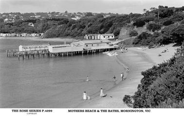 This shows a sweeping view of the beach with the Baths in the centre of the photo. There are several people on the beach, with a mother and small child in the foreground centre. It appears to be a warm sunny day. There are lots of bathing huts shown. There are lots of houses on the crest of the embankment. The waters of Port Philip Bay are still and clear. There are a row of Kayaks shown on the beach, and several just on the water’s edge.