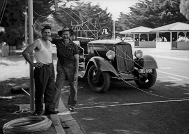 In the foreground two men are standing beside an open  roadster, registration 81.056. The bare headed man on the left is Max Pender of the C.F.A., the identity of the man on the right in army hat is unknown. A fire bell, fire tower and two rotundas are in the background in front of a line of trees beside the training track. 