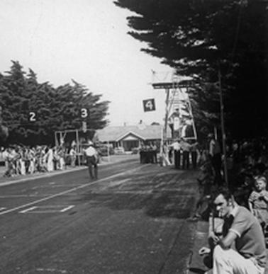 A portable tower is positioned on the right of a broad asphalted track. A man is standing part way up a ladder which provides access to a high platform. Officials are gathered at the foot of the tower. Onlookers line the sides of the track. Mornington House is in the background.