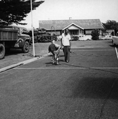 A macadamised track in the foreground with a house signed ‘Mornington House’ in background. An old fire engine is to the left of the running track and a fire hose snakes from the engine on to the track. A man stands on the track and another in running shorts is bent over in front of the hose. 