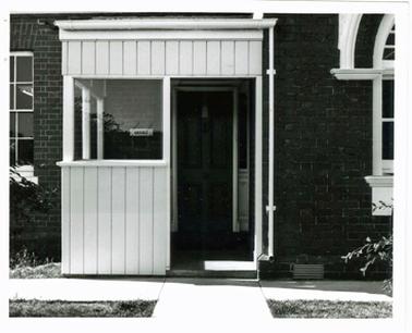 This shows the front porch of the Police Station in Mornington. It is a white timber enclosed porch, and there is a sign beside the front door “Office”. The building is brick, and to the right of the porch is an arched window. To the right of the door is a sash window.