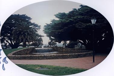 This is a frontal view of a fountain at one end of a circular brown stone wall. A low jet of water falls on to a platform creating the effect of a waterfall. The remainder of the enclosed ground is covered in shallow water. There is no visible outlet for the water. There is a cypress tree on either side between the stone wall and the sea. A white poat and rail fence stands between the edge of the park and the sea which appears in the background. Several cars are parked near the fence.