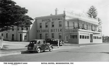 This shows the Royal Hotel which is a two storey grand building with verandahs to both levels. It is a brick building which has been rendered white. The ground floor veranda is closed in. There are decorative urns to the front roof, and a pediment in the centre front showing the name of the hotel. There is a sign on the side of the hotel with the name and “Victoria Bitter”. There are two black cars shown in the foreground. There is a large Pine tree to the right of the hotel.
