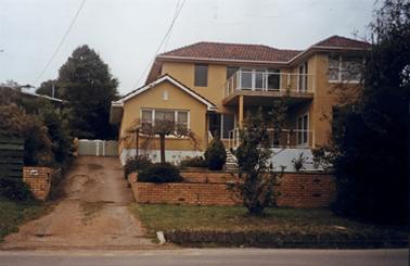 A two story house in sand coloured rendering stands in a small garden behind a terraced low cream brick fence. It has a terrace in front defined by a transparent balustrade, a flight of steps and a low white wall. The facade has several white framed windows and a French window on to the terrace. The second story covers most of the ground floor and includes a balcony over the front entrance with a transparent balustrade. There is a gravel drive on the left with a strip of garden along the side fence and a sparsely vegetated front garden.