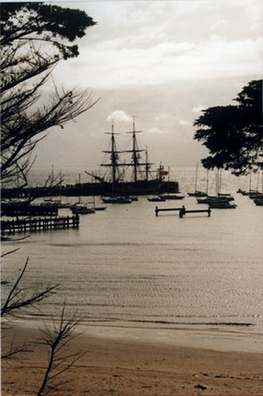 View from a beach across calm water to a pier. There is a large yacht with bare rigging beside the pier and  several small yachts in the foreground. The pier is framed by trees at the edges of the scene.