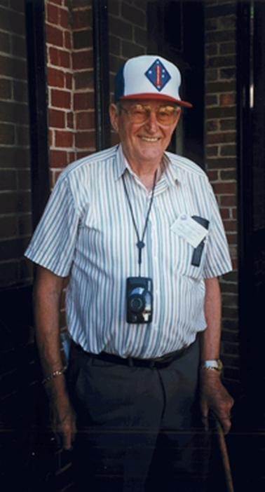 A bespectacled, smiling man of about 70 years is standing in front of a brick wall. He is dressed casually in a red, blue and white striped shirt with short sleeves and an open neck with a peaked cap in the same colours. There is a diamond shaped badge in front with white stars in the shape of the Southern Cross on a blue background. A badge is pinned to his shirt pocket and there is a camera hanging from a cord around his neck. He appears to hold a walking stick in his left hand.