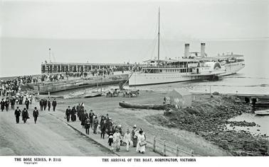 This photograph shows lots of people making their way off the Pier having just arrived on the steam boat Hygea. The Hygea is a paddle steamer with two funnels. The people are wearing Victorian dress and some are carrying large bags suggesting they may be on holiday. There are two motor vehicles shown in the centre of the picture, and s couple of fishing boats at the waters edge.