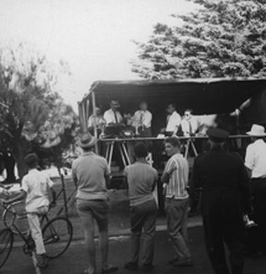 A platform with trestle tables at the front is covered by a tarpaulin. Several men in shirtsleeves and ties are behind the tables and a shield with a metal trophy beside it. Several onlookers are standing in front and there are trees behind the stand.  