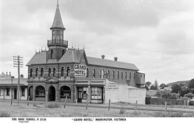 This image shows the Grand Hotel in it’s original state. It is an impressive two storey brick building with a tower. There is a central arched doorway, with an arched window  on either side. To the left as we face the hotel, there is a third arch which was the original stables, this has a metal gate. There is a large sign on the right side of the Hotel “Grand Hotel, L. Harrison”. There is a private dwelling to the left of the hotel, and two shops to the right. The business name on one shop is J. Livingstone & Co, Auctioneers and Surveyors. On the front windows are signs saying Billiards Room and Bar. The roof is steeply pitched and slate. 